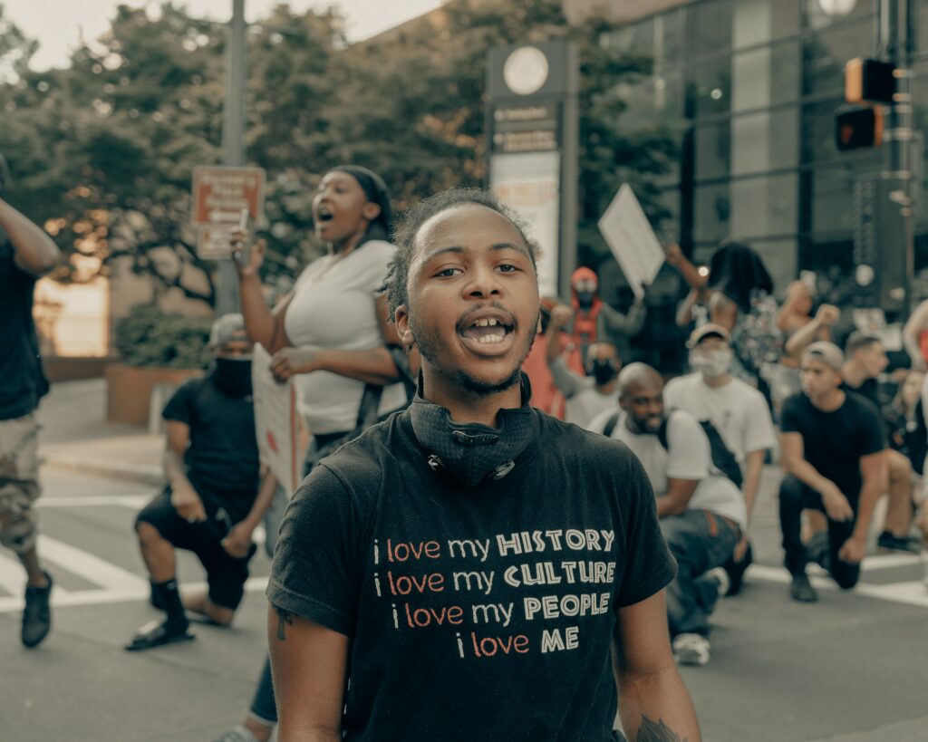 Black activist at a protest, wearing a t shirt that says "i love my history, i love my culture, i love my people, i love me"