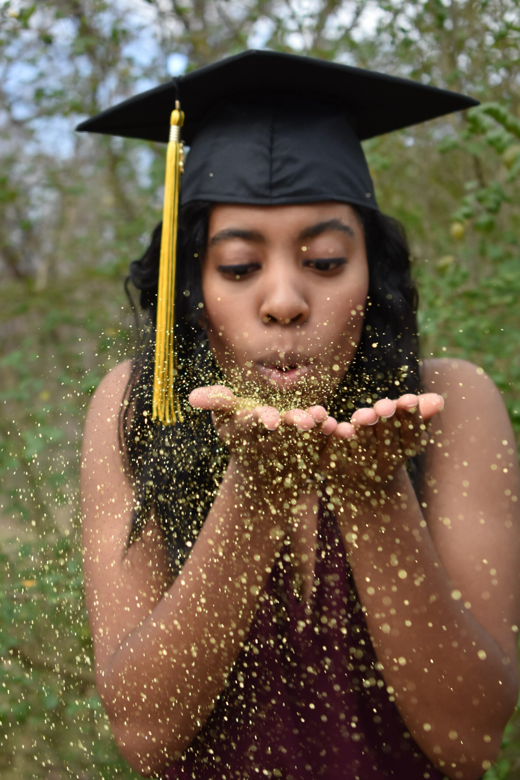 A black woman wearing a graduation cap celebrating by blowing glitter from her hands into the air.